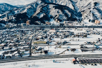 A sprawling town covered in snow lies at the base of rugged, forested mountains. Numerous buildings, both tall and short, are scattered throughout the area, while roads crisscross the urban landscape. The mountainous backdrop is heavily snow-laden, with dark trees creating a striking contrast. Several structures stand prominently near the mountains, suggesting a mix of residential and commercial use.