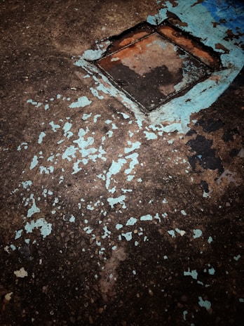 A concrete surface with peeling paint reveals patches of underlying material, with a square metallic or concrete cover embedded. The paint is chipping away, particularly around the edges of the square section, exposing a rusty color beneath the blue paint.