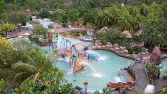A vibrant water park featuring multiple pools, slides, and water play structures surrounded by lush greenery and palm trees. The area is filled with colorful slides and cascading water features, creating a lively atmosphere.