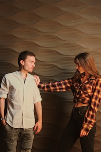 A man and a woman stand against a stylish textured wall. The man wears a light-colored shirt and jeans, while the woman is dressed in a plaid shirt and dark jeans. She is reaching out to touch the man’s shoulder, creating a slightly tense interaction.