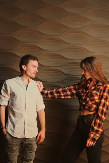 A man and a woman stand against a stylish textured wall. The man wears a light-colored shirt and jeans, while the woman is dressed in a plaid shirt and dark jeans. She is reaching out to touch the man’s shoulder, creating a slightly tense interaction.