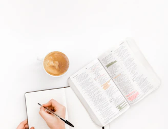 A close-up of hands writing in notebooks beside a cup of coffee.
