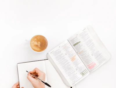 A close-up of hands writing in notebooks beside a cup of coffee.