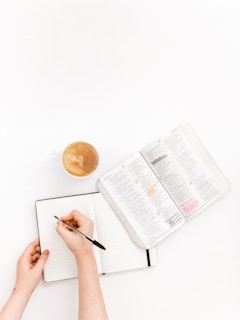 A person's hand writing in a lined notebook with an open book nearby, possibly a textbook or reference material. There is a cup of coffee placed to the left side, suggesting a study or work environment.