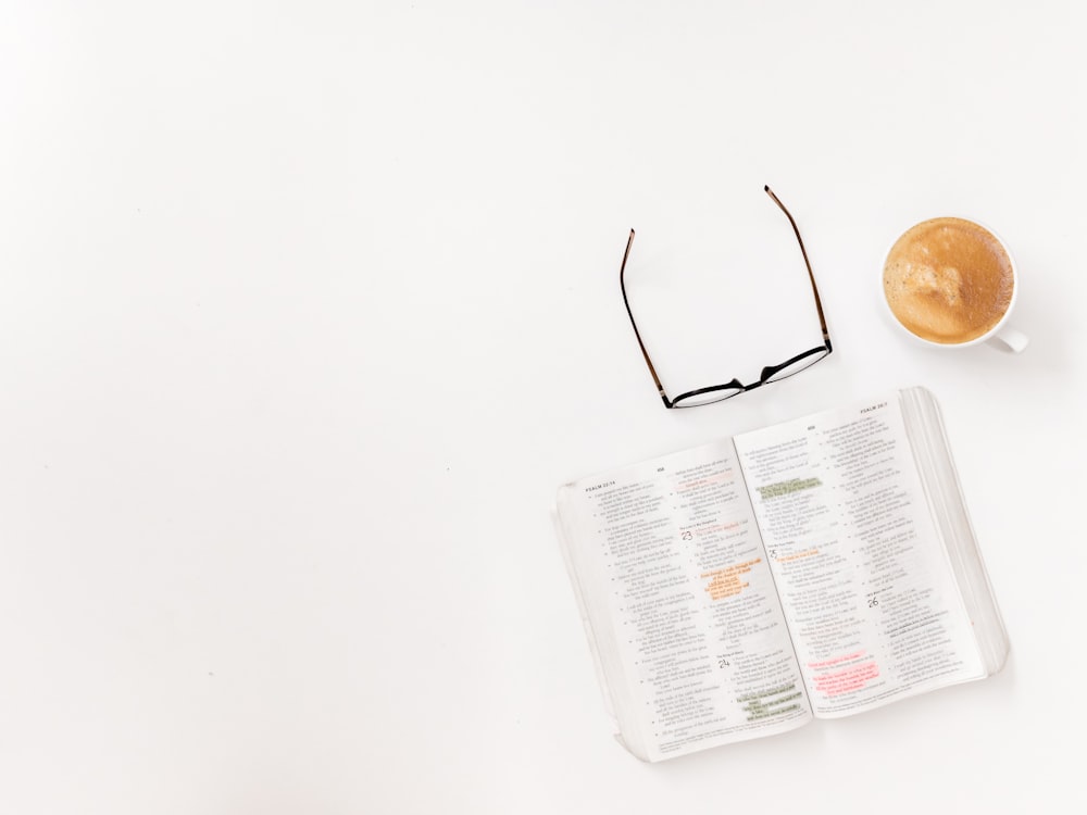 eyeglasses with black frames beside cappuccino glass and printed book on white surface