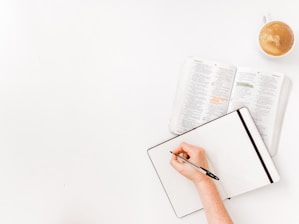 A hand writing in a large, open notebook with blank, lined pages. Next to the notebook is an open book with text highlighted in various colors. In the top right corner, there is a cup of coffee with foam on top.