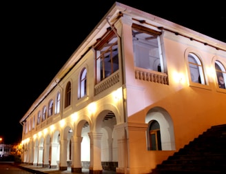 A well-lit, two-story building with arched windows and columns. The architecture features classic elements with white walls and stone accents. Decorative lighting enhances the structure's elegance during nighttime.