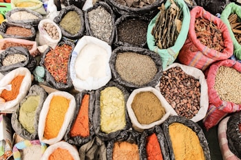A colorful and diverse array of spices, herbs, grains, and dried goods are displayed in open bags. Each bag contains a different item, creating a mosaic of textures and colors. Some of the items appear finely ground, while others are whole or coarsely chopped, with earthy tones dominating the scene.