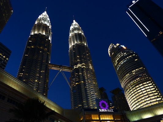 Tall, illuminated skyscrapers stand against the night sky, joined by a skybridge. The foreground features a building with signs for Suria KLCC, adding to the urban landscape.