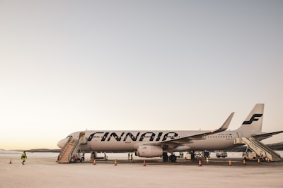An airplane with the FINNAIR logo is parked on the tarmac. Portable stairs are attached for passenger boarding, and a few people in high-visibility vests are present around the aircraft. The scene appears to be during the day with a clear sky, and traffic cones are placed around the area.