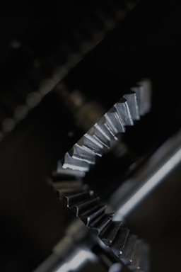 Two interlocking metal gears with detailed teeth patterns are positioned against a dark background. The gears are metallic and reflect light, emphasizing their textured surfaces.