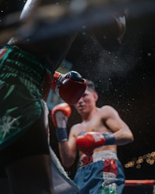 Two boxers engaged in a boxing match, with one boxer landing a punch. The focus is on the movement and dynamics of the fight, highlighted by the action of the red and blue boxing gloves. The background is blurred, emphasizing the intensity and physicality of the scene.