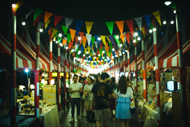 A vibrant night market with colorful triangular flags hanging overhead and illuminated by string lights. Stalls are lined on both sides displaying various goods, while people casually stroll and browse. The atmosphere is lively and bustling.