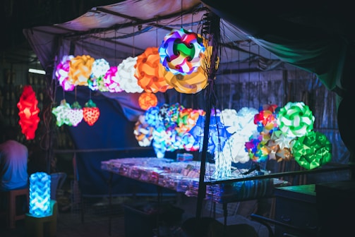 Various colorful, intricately designed lamps are displayed hanging in a market setting. The lamps have geometric and abstract patterns, casting a vivid array of colors in the surrounding dark environment. A table beneath the lamps holds additional items, possibly more lamps or related products.