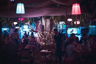 A dimly lit outdoor restaurant or bar with groups of people sitting at tables enjoying food and drinks. The ambiance is set by colorful hanging lanterns and string lights draped around trees and structures. A waiter is seen attending to a table. The atmosphere appears lively and social, with most patrons engaged in conversation.