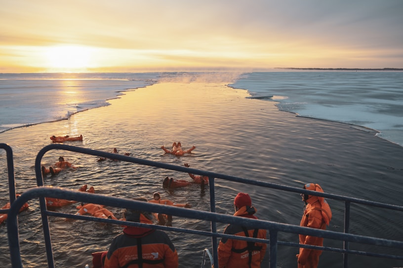 A group of people wearing orange survival suits are seen swimming in icy waters, with an icebreaker nearby. The ice-covered surface contrasts with the open water, and a golden sunset illuminates the scene, casting warm reflections on the water.