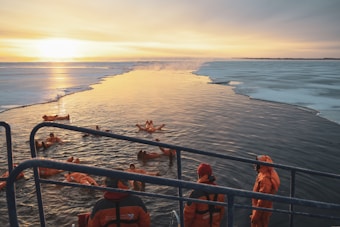 A group of people wearing orange survival suits are seen swimming in icy waters, with an icebreaker nearby. The ice-covered surface contrasts with the open water, and a golden sunset illuminates the scene, casting warm reflections on the water.