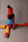 A creatively styled abstract figure is composed of various colorful, textured materials. The figure appears to be seated on a neutral gray background. Bright colors such as red, orange, yellow, blue, and pink are prominent in the wrapped materials.