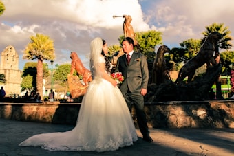 A bride in a white wedding gown and a groom in a gray suit stand facing each other in a sunlit outdoor setting. Behind them is a fountain featuring statues of large cats. The scene is surrounded by palm trees and historical architecture, with a vibrant blue sky and fluffy clouds overhead.
