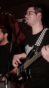 A musician is smiling while singing into a microphone. He is playing a bass guitar with a strap that has a black-and-white checkered pattern. Another person is partially visible in the background. The lighting is dim, suggesting a cozy or intimate performance setting.