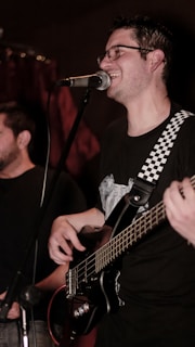A musician is smiling while singing into a microphone. He is playing a bass guitar with a strap that has a black-and-white checkered pattern. Another person is partially visible in the background. The lighting is dim, suggesting a cozy or intimate performance setting.
