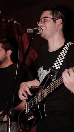 A musician is smiling while singing into a microphone. He is playing a bass guitar with a strap that has a black-and-white checkered pattern. Another person is partially visible in the background. The lighting is dim, suggesting a cozy or intimate performance setting.