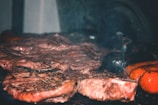 A close-up view of a grill with various types of meat being cooked, including steaks and sausages. The meat appears juicy and well-seasoned, with visible grill marks and a hint of smoke rising. The scene is likely from a barbecue setting.