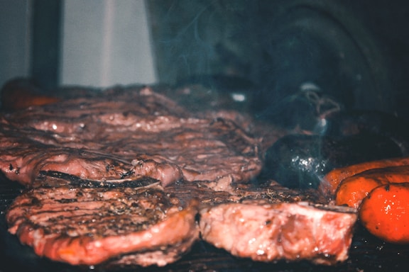 A close-up view of a grill with various types of meat being cooked, including steaks and sausages. The meat appears juicy and well-seasoned, with visible grill marks and a hint of smoke rising. The scene is likely from a barbecue setting.