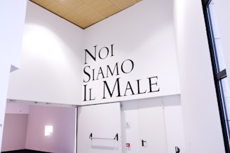 A minimalist interior space with high white walls featuring the phrase 'Noi Siamo Il Male' in bold black letters. The area has a wooden ceiling and a minimalistic modern design. There is a doorway with no door and a visible disability access sign.
