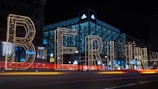 Large illuminated letters spell out 'Berlin' against the backdrop of a city street at night. The scene is vibrant with lights outlining the letters and colorful streaks created by passing cars. Behind the letters, an Adidas store stands brightly lit, adding to the urban atmosphere of a bustling night.