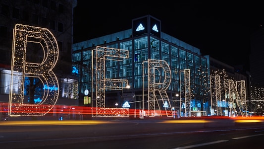 Large illuminated letters spell out 'Berlin' against the backdrop of a city street at night. The scene is vibrant with lights outlining the letters and colorful streaks created by passing cars. Behind the letters, an Adidas store stands brightly lit, adding to the urban atmosphere of a bustling night.