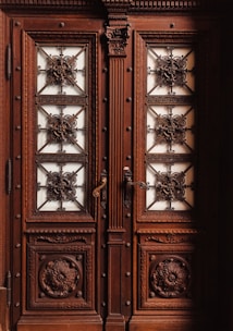A pair of ornate double doors made of dark wood featuring intricate carvings with floral and geometric patterns. The panels are adorned with metal grillwork that forms elaborate, decorative designs. The door handles are metal and intricately designed as well.