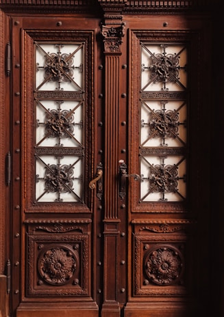 A pair of ornate double doors made of dark wood featuring intricate carvings with floral and geometric patterns. The panels are adorned with metal grillwork that forms elaborate, decorative designs. The door handles are metal and intricately designed as well.