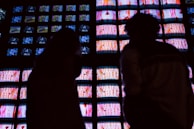 A wall of numerous television screens displays colorful images, with a silhouette of two people standing in front of it. The screens emit bright hues, creating a stark contrast with the dark silhouettes.