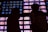 A wall of numerous television screens displays colorful images, with a silhouette of two people standing in front of it. The screens emit bright hues, creating a stark contrast with the dark silhouettes.