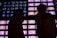 A wall of numerous television screens displays colorful images, with a silhouette of two people standing in front of it. The screens emit bright hues, creating a stark contrast with the dark silhouettes.