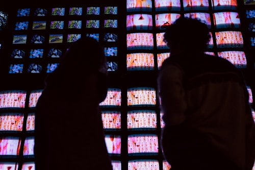A wall of numerous television screens displays colorful images, with a silhouette of two people standing in front of it. The screens emit bright hues, creating a stark contrast with the dark silhouettes.