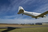 A classic propeller-driven airplane is flying low over an airfield with clear skies in the background. The ground features runway markings and a grassy area, with a hangar and other buildings visible in the distance. The plane is white, and some details like windows and the propeller are discernible.