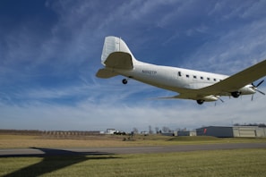 A classic propeller-driven airplane is flying low over an airfield with clear skies in the background. The ground features runway markings and a grassy area, with a hangar and other buildings visible in the distance. The plane is white, and some details like windows and the propeller are discernible.