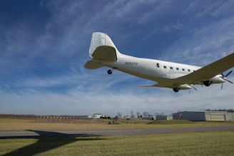 A classic propeller-driven airplane is flying low over an airfield with clear skies in the background. The ground features runway markings and a grassy area, with a hangar and other buildings visible in the distance. The plane is white, and some details like windows and the propeller are discernible.