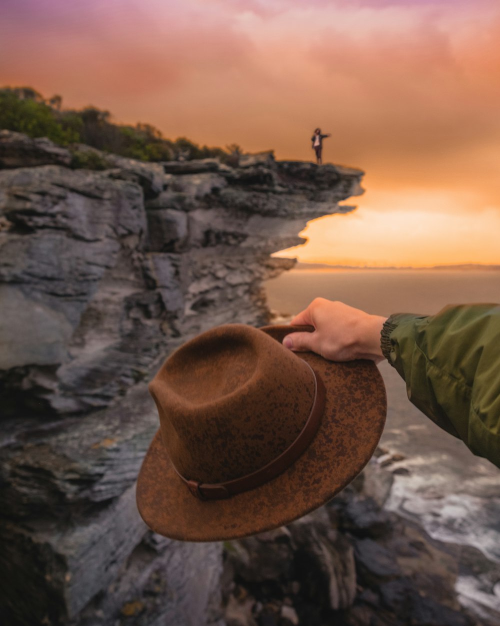 man about to throw his hat at the sea in front of man waving at the edge of the cliff