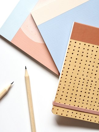 A flat lay of three notebooks in different pastel colors and designs. Two wooden pencils with sharpened tips are placed alongside the notebooks, creating a neat and organized arrangement on a clean white surface.