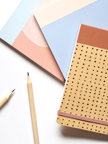 A flat lay of three notebooks in different pastel colors and designs. Two wooden pencils with sharpened tips are placed alongside the notebooks, creating a neat and organized arrangement on a clean white surface.