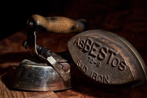 An antique flat iron with a wooden handle and metallic body is shown. Engraved on the base of the iron is the word 'ASBESTOS' along with other text. The iron's surface is worn, indicating its age and historical use.