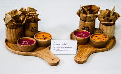 Wooden serving boards hold bowls of colorful hummus and containers filled with crisp, folded lavash bread. Small bowls contain beetroot and red pepper hummus, distinguished by their vibrant pink and orange colors. A card in front provides a hand-written description of the flavors.