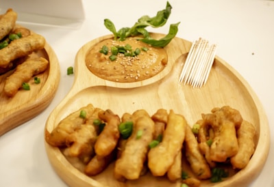 A wooden serving platter holds a pile of deep-fried snacks garnished with chopped green onions. Next to the snacks is a small container of dipping sauce topped with sesame seeds and a sprig of fresh basil. A set of toothpicks is placed beside the sauce for serving.