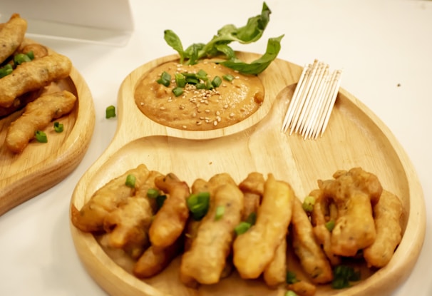 A wooden serving platter holds a pile of deep-fried snacks garnished with chopped green onions. Next to the snacks is a small container of dipping sauce topped with sesame seeds and a sprig of fresh basil. A set of toothpicks is placed beside the sauce for serving.