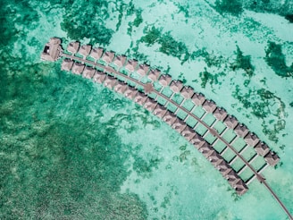 Aerial view of a series of overwater bungalows forming a curved line in a turquoise lagoon, showcasing clear waters with patches of darker green coral beneath.