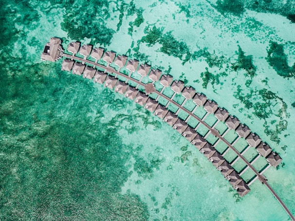 Aerial view of a series of overwater bungalows forming a curved line in a turquoise lagoon, showcasing clear waters with patches of darker green coral beneath.