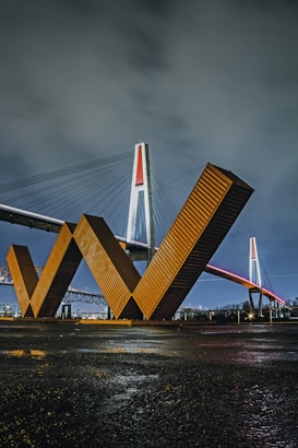 A bridge with a distinctive orange and red lighting design spans across the image, with cables and a tall tower structure in view. The foreground features large geometric metal structures resembling the letter 'W', set against a cloudy night sky.